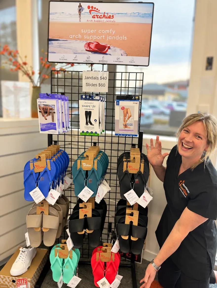 A smiling member of the Masterton Foot Clinic team waving beside the Archies arch support jandals and slides display stand in the clinic